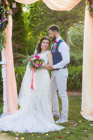 New husband and wife posing under textile arch at their wedding ceremony in the park with pink accessories